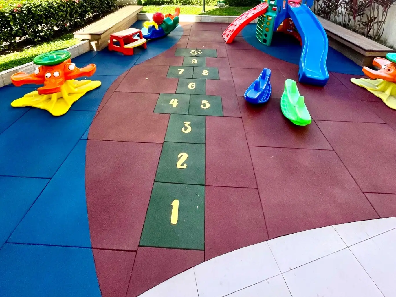 Área de brincar com piso de borracha em placas azuis, vermelhas e verdes com amarelinha